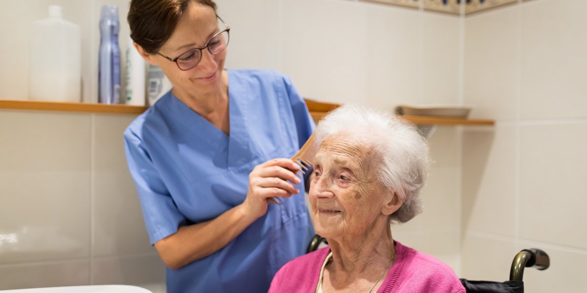 Ein Pflegerin kämmt einer alten Frau die Haare. Sie sind in einem Badezimmer.
