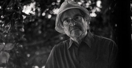 Älterer Mann mit Strohhut und Brille unter einem Baum.