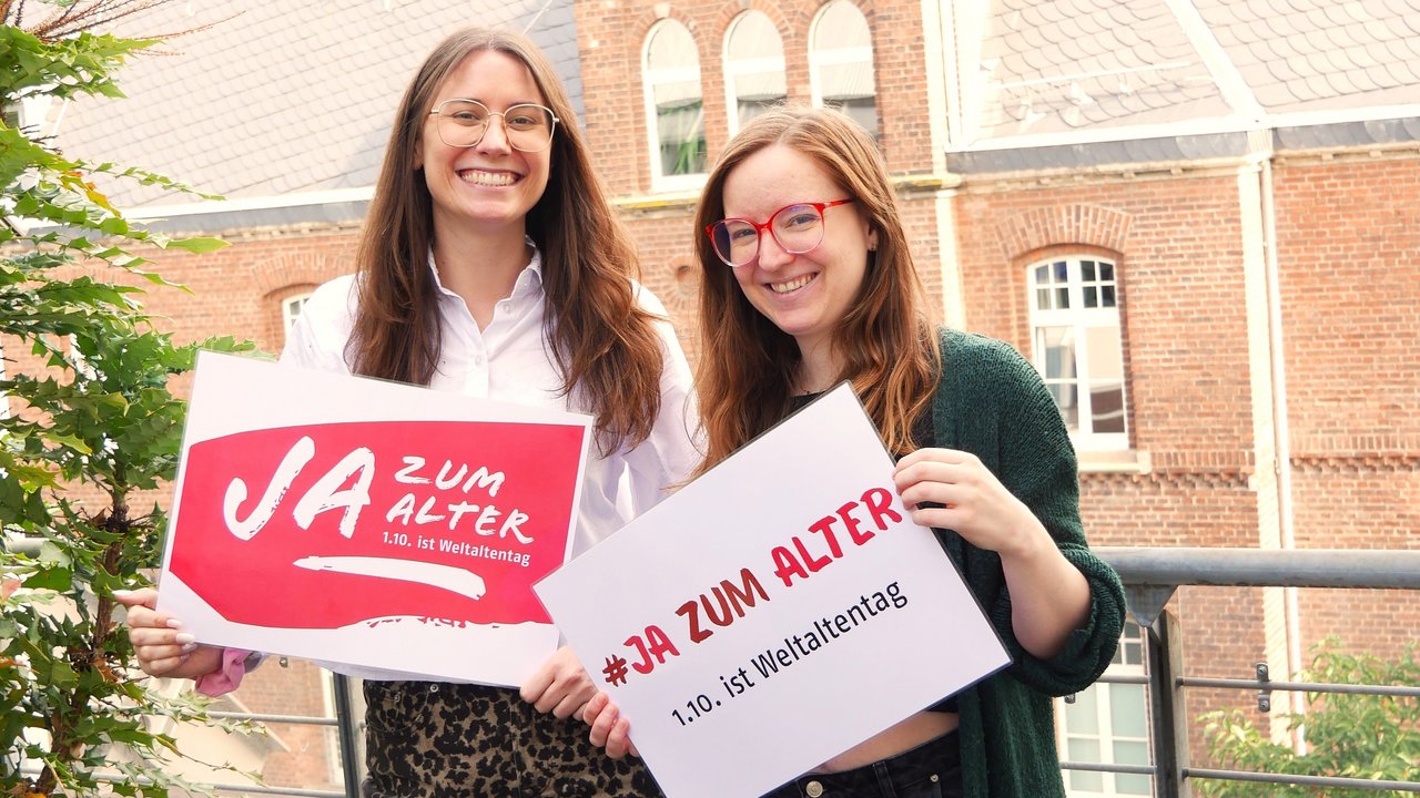 Zwei junge Frauen stehen auf einer Terrasse. Sie halten jeweils ein Schilder mit der Aufschrift "JA zum Alter" in der Hand. Klick öffnet eine vergrößerte Ansicht.