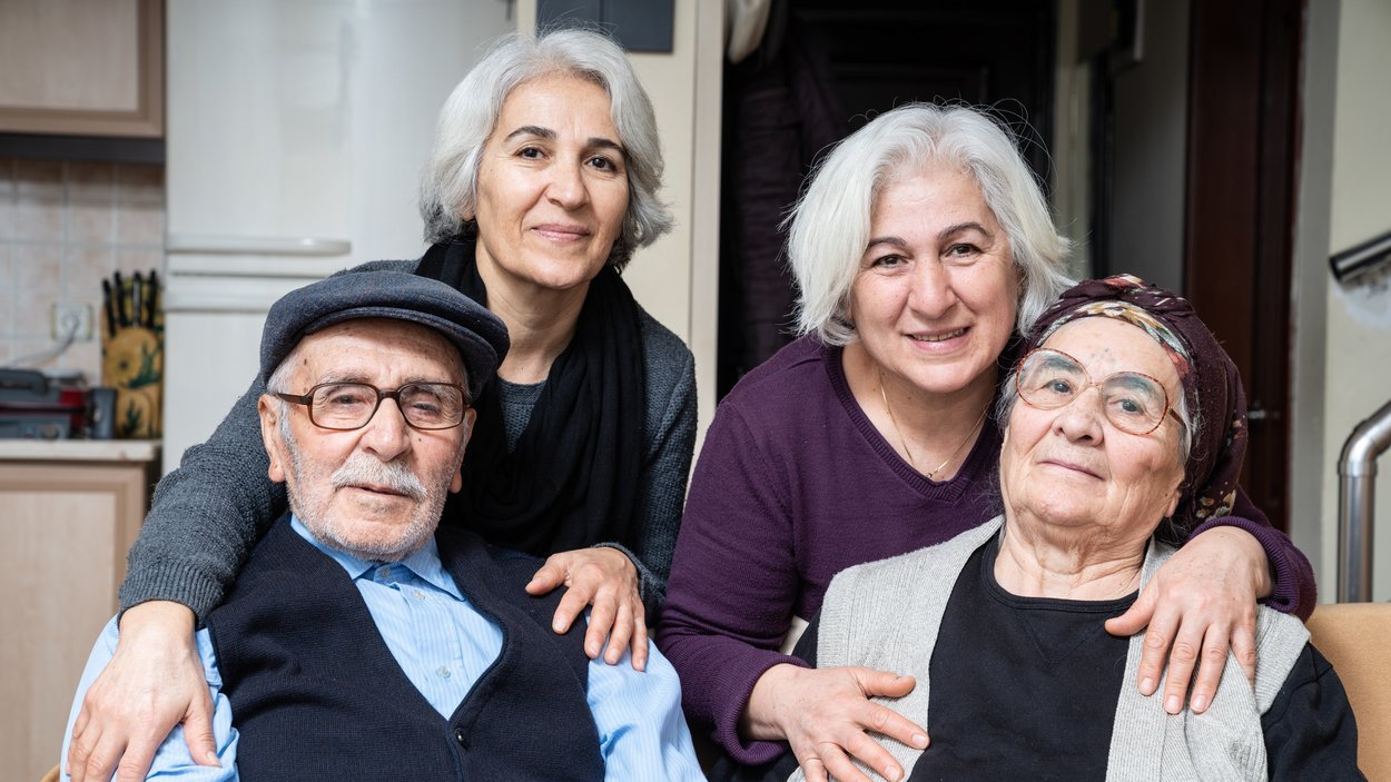 ©selimaksan_istock.com Ein älteres Paar sitzend, dahinter stehen zwei Frauen mitleren Alters, die ihre Hände auf die Schultern des Paares gelegt haben.