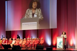Blick auf eine Bühne: Im Vordergrund sieht man ältere Menschen auf Stühlen sitzen. Sie tragen Kleidung in Rottönen. Hinter ihnen ist eine Musiband zu sehen. Über ihnen ist auf einer großen Leinwand eine ältere Frau mit krausem Haar zu sehen. Sie steht auf der Bühne an einem Pult, um eine Rede zu halten.