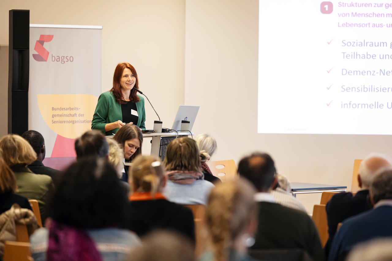 ©BAGSO_Seng The speaker Ute Hauser, a young woman with long red hair, is standing at a lectern. She is wearing a black T-shirt and a green blazer over it. A BAGSO banner can be seen behind her. Participants in the conference are sitting in the foreground. Klick öffnet eine vergrößerte Ansicht.