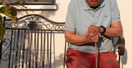 Älterer Mann mit Stock in der Hand sitzt auf einer Bank vorm Haus und zwinkert.