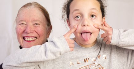 Ältere Frau lacht, neben ihr streckt Enkelin die Zunge heraus und zieht die Augen nach unten