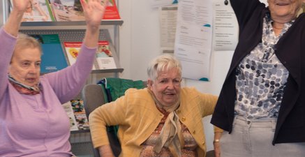 3 Hochaltrige tanzen. Rechts im Stehen. In der Mitte stützt sich im Sitzen ab, links im Sitzen& Hände über Kopf