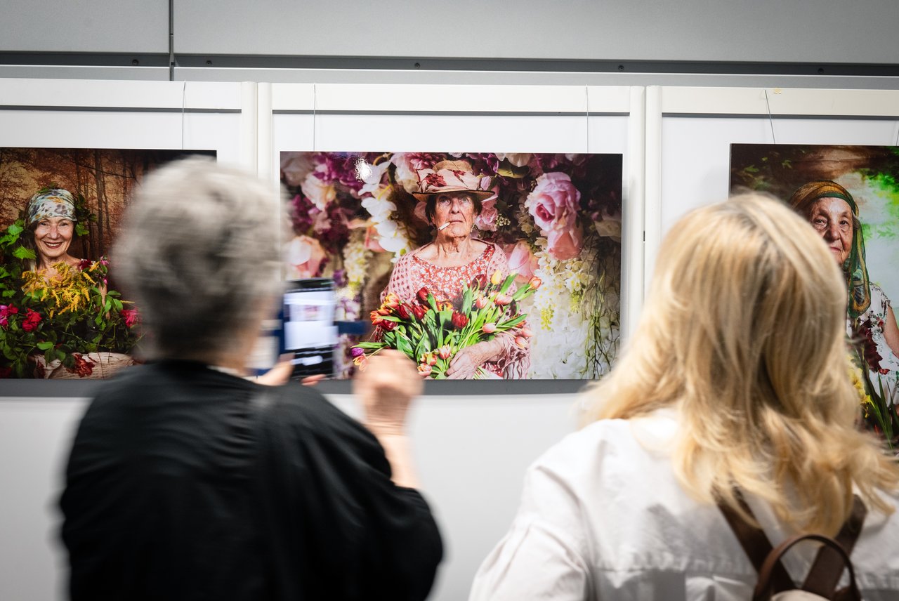 Zwei Frauen stehen vor einer Fotografie. Darauf ist eine alte Frau mit Hut zu sehen. Sie trägt ein rötlich, mit Blumen bedrucktes Kleid. Im Mund hat sie eine Zigarette. In den Händen eine großen Strauß roter Tulpen. Sie ist umrahmt von einem Bild, auf dem weiße und rosa Rosen in Nahaufnahme zu sehen sind. Klick öffnet eine vergrößerte Ansicht.