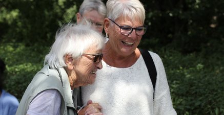 Zwei ältere Damen halten sich vertraut lachend an den Händen. Im Hintergrund ist eine grüne Hecke.