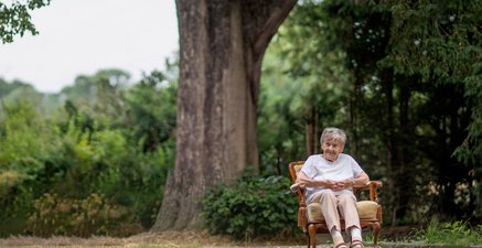 Eine 100 jährige Frau sitzt auf einem Sessel im Garten. Hinter ihr steht eine große Linde.