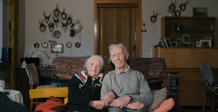 Ein Ehepaar sitzt zusammen auf einem Sofa in ihrem Wohnzimmer und schaut in die Kamera. Sie wirken fröhlich.