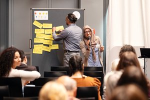 Ein Workhopraum: Vorne eine Moderationswand mit einigen beschrifteten gelben Karten. Ein Mann steht mit dem Rücken zum Publikum und pint gerade eine Karte an. Dvor steht eine junge Frau, mit beigem Kopftuch und einer Brille. Sie lächelt freundlich und spricht in ein Mikrofon.