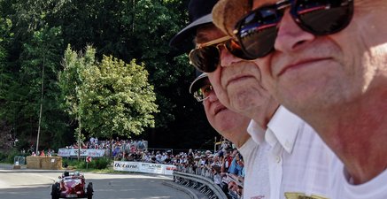 Drei alte Drillingsbrüder am Streckenrand einer Straße, auf der ein Oldtimer-Rennwagen fährt.
