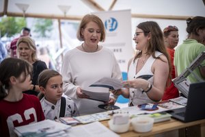 Eine Frau mittleren Alter steht an einem Messestand. Neben ihr zwei Kinder und auf der anderen Frau eine junge Frau, die ihr eine Broschüre zeigt.