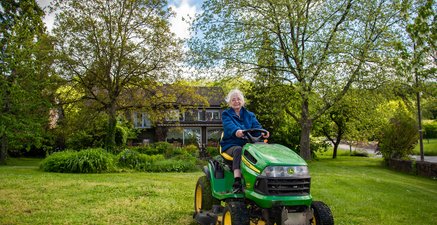 Ältere Dame sitzt auf Rasenmähertrecker in einem Garten und lacht in die Kamera