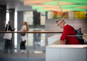 Eine ältere Frau steht auf einem Balkon in einer Messehalle. Sie trägt einen roten Pullover und hat einen Rucksack an. Sie schaut in die untere Etage. Im verschwommenen Hintergrund sieht man zwei Frauen, die sich unterhalten.