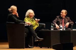 Bühn: Eine alte Frau mit grüner Bluse, Gesundheitswissenschaftlerin Annelie Keil und ein alter Mann, der ehemalige Minister Franz Müntefering, sitzen auf der Bühne in scharzen Sesseln. Frau Keil spricht gerade in ein Mikrofon. Bei ihnen sitzt eine blonde Frau, die Moderatorin.