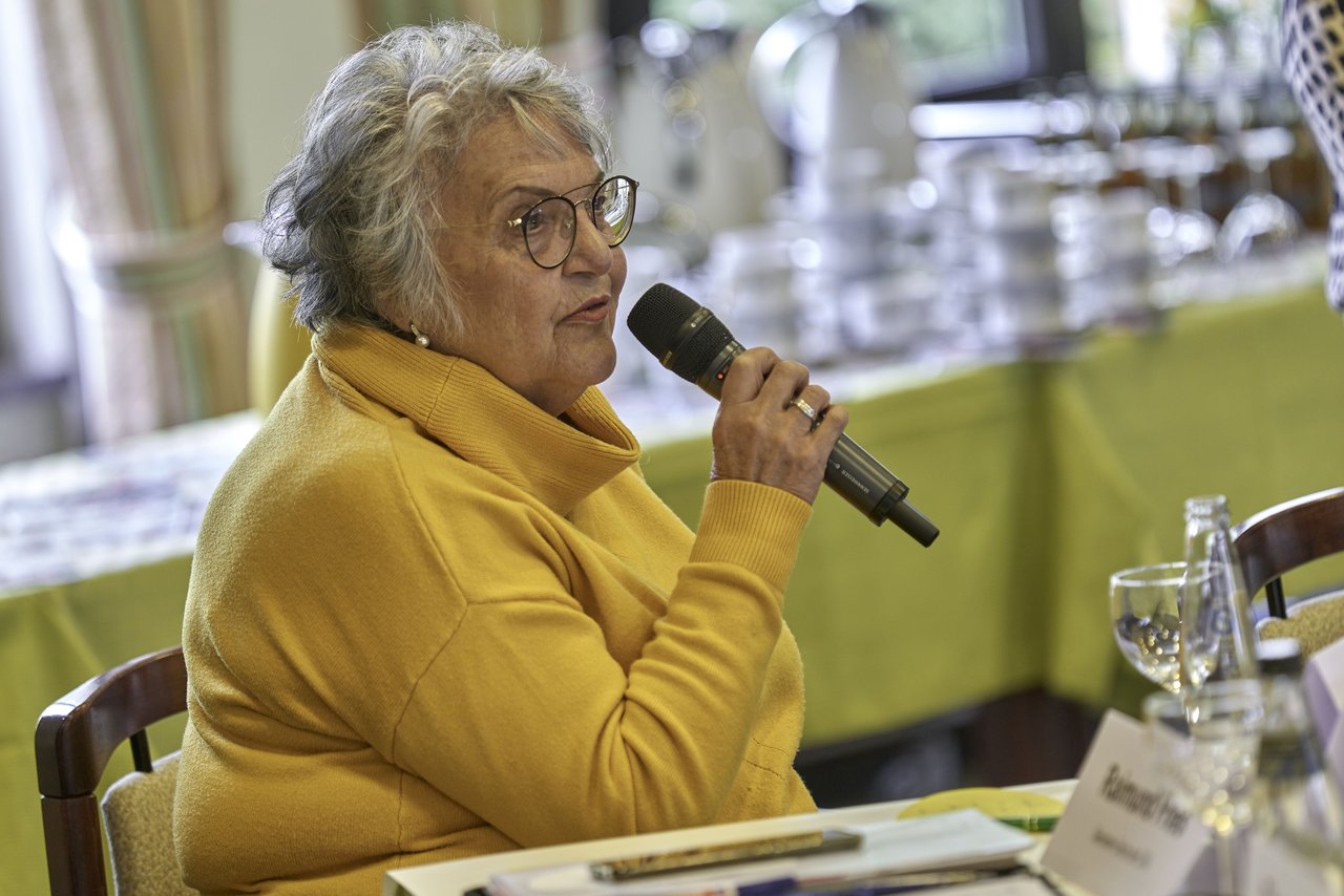 Eine ältere Frau sitzt an einem Tisch und spricht in ein Mikrofon.