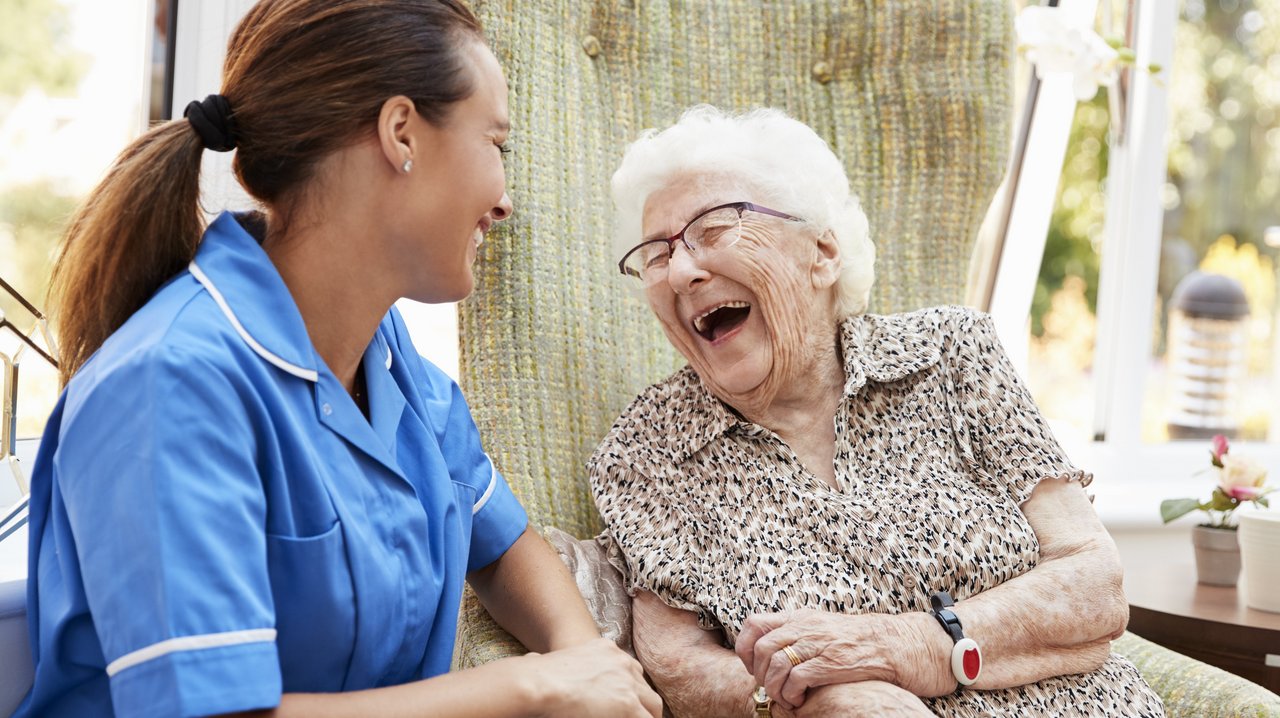 Pflegerin sitzt bei herzhaft lachender alter Frau.