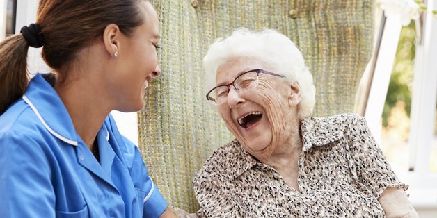 Eine Pflegerin sitzt neben einer alten Frau, die herzhaft lacht.