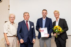 Eine Frau und drei Männer stehen auf der Bühne. Ein junger Mann hält ein Zertifikat in die Kamera, ein zweiter junger Mann hat einen Blumenstrauß in der Hand.