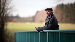 Ein alter Mann steht auf einer Brücke. Im Hintergrund sieht man Bäume und ein Gebäude.