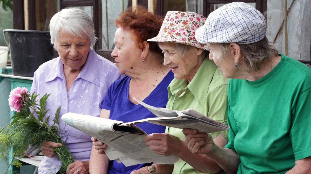 Vier ältere russische Frauen sitzen vor einem Haus auf einer Bank und lesen Zeitung