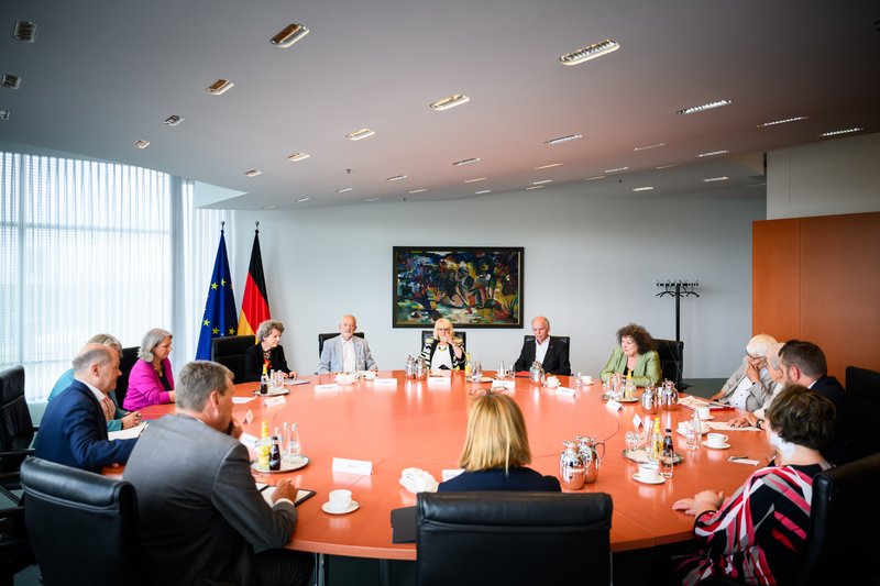 14 Personen sitzen um einen runden Konferenztisch aus Kirschbaumholz. Im Hintergrund sieht man die Deutschland- und Europafahne und ein abstraktes Gemälde. 