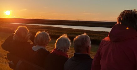 Fünf Freunde sitzen auf einer Bank am Deich und genießen den Sonnenuntergang.