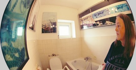 Fischaugen-Aufnahme einer Frau, die nachdenklich in ihr Badezimmer mit Toilettenaufsatz und Badewannenbrett schaut 