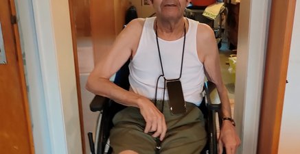 Ein älterer Mann sitzt im Unterhemd im Rollstuhl. Auf dem Kopf hat er einen Damenhut. Er lächelt vorsichtig.