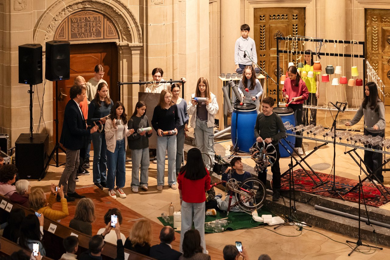 Eine Szene in der Kirche. Kinder und Jugendliche spielen auf selbst gebauten Instrumenten. Zum Beispiel auf umgedrehten bunten Blumentöpfen, die an Seilen Hängen. Zwei Mädchen trommeln auf Industriefässer. Ein Junge hat Schlappen in der Hand und schlägt damit auf graue Wasserrohre, die in einem Tisch eingebaut sind. Ein anderes Mädchen schlägt mit Schlegeln auf Plastikflaschen, die auf einen tischähnlichem Rahmen montiert sind. Auf dem Boden ist ein umgedrehtes Fahrrad zu sehen, vor dem zwei Jungen sitzen. Klick öffnet eine vergrößerte Ansicht.