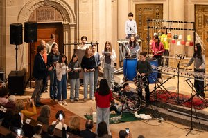 Eine Szene in der Kirche. Kinder und Jugendliche spielen auf selbst gebauten Instrumenten. Zum Beispiel auf umgedrehten bunten Blumentöpfen, die an Seilen Hängen. Zwei Mädchen trommeln auf Industriefässer. Ein Junge hat Schlappen in der Hand und schlägt damit auf graue Wasserrohre, die in einem Tisch eingebaut sind. Ein anderes Mädchen schlägt mit Schlegeln auf Plastikflaschen, die auf einen tischähnlichem Rahmen montiert sind. Auf dem Boden ist ein umgedrehtes Fahrrad zu sehen, vor dem zwei Jungen sitzen.