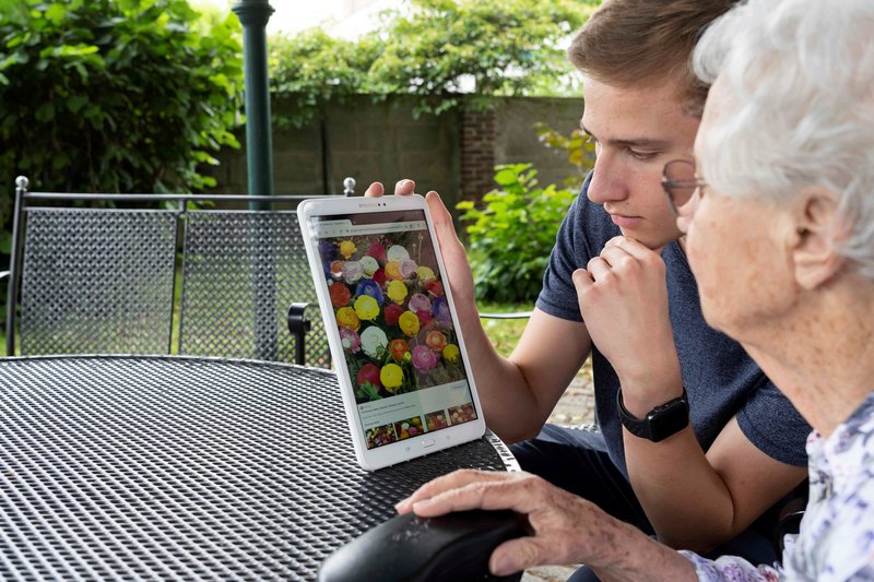 Ein junger Mann und eine alte Frau sitzen im Garten am Tisch und schauen auf ein Tablet.