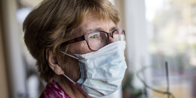 Eine ältere Frau schaut mit Mund-Nasen-Schutz aus dem Fenster.