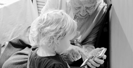 Eine 93-Jährige sitzt auf einem Stuhl und hält ein Handy in der Hand. Sie zeigt ihrer Ur-Enkelin ein Familienfoto.