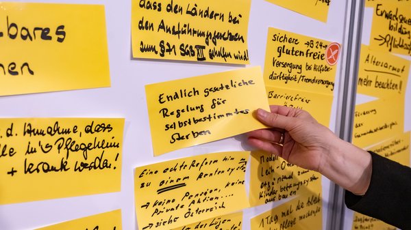 Eine Pinwand, auf der beschriftete gelbe Zettel kleben. Eine Notiz wird gerade von einer Hand angeheftet.