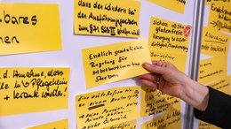 Eine Pinwand, auf der beschriftete gelbe Zettel kleben. Eine Notiz wird gerade von einer Hand angeheftet.