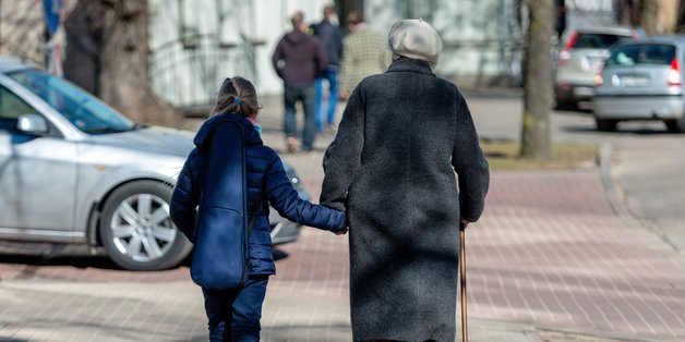 Rückenansicht einer älteren Frau mit Gehstock und einem kleinen Mädchen, die Hand in Hand auf einem Gehsteig gehen