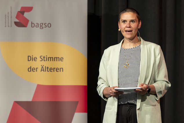 Eine junge Frau steht auf der Bühne. Sie hat Karteikarten in der Hand und spricht gerade in ein Headset. Klick öffnet eine vergrößerte Ansicht.