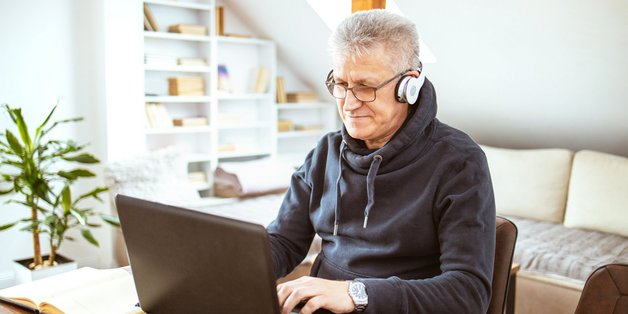 Älterer Mann mit Headset sitzt in seinem Wohzimmer vor einem Laptop