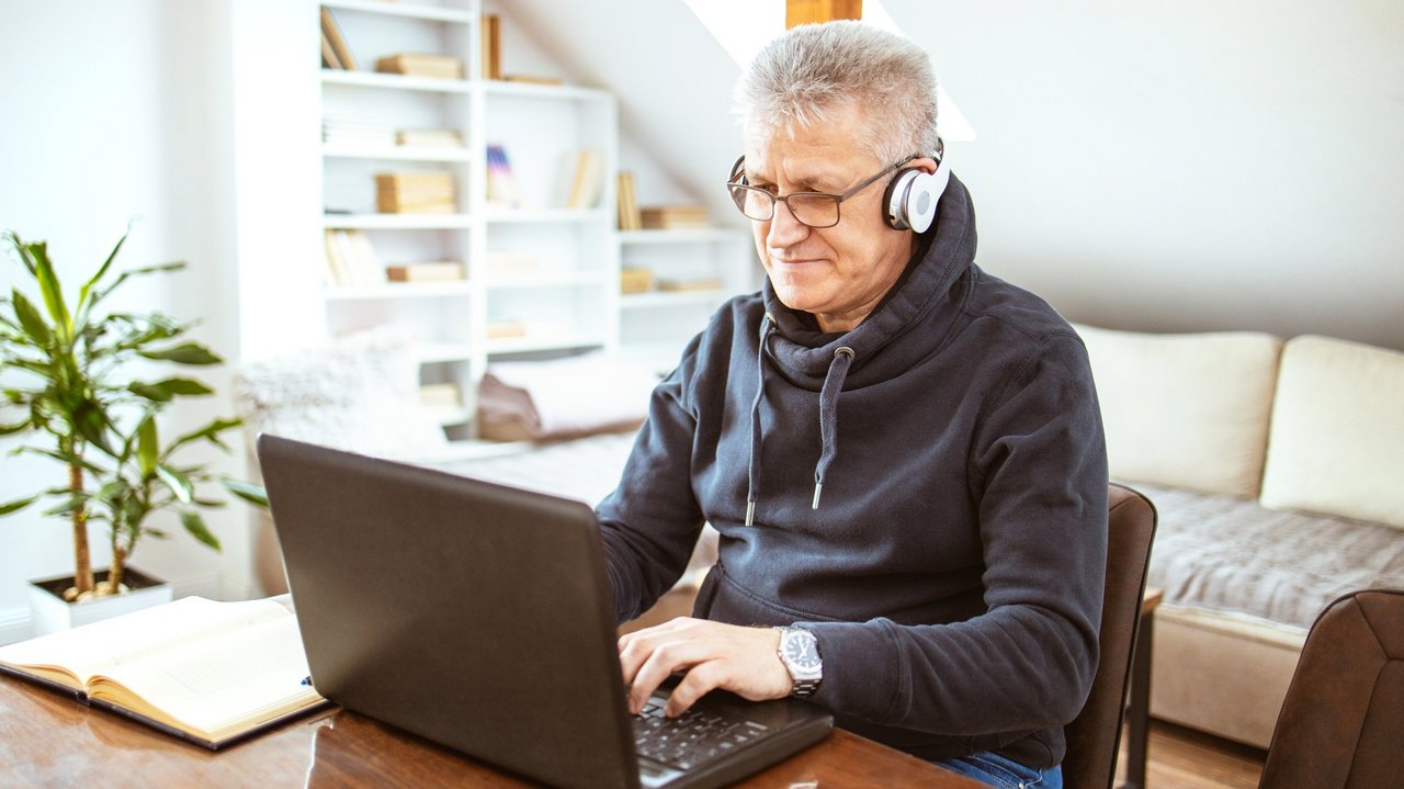 ©lka84_istock.de Älterer Mann mit Headset sitzt in seinem Wohzimmer vor einem Laptop