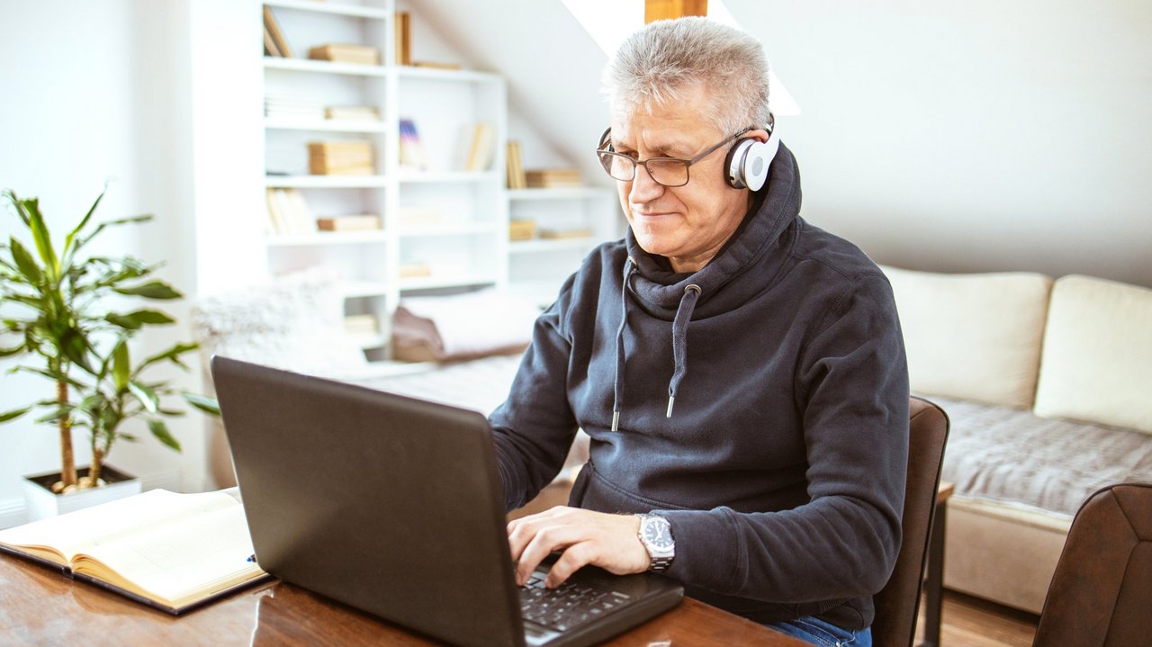 Älterer Mann mit Headset sitzt in seinem Wohzimmer vor einem Laptop