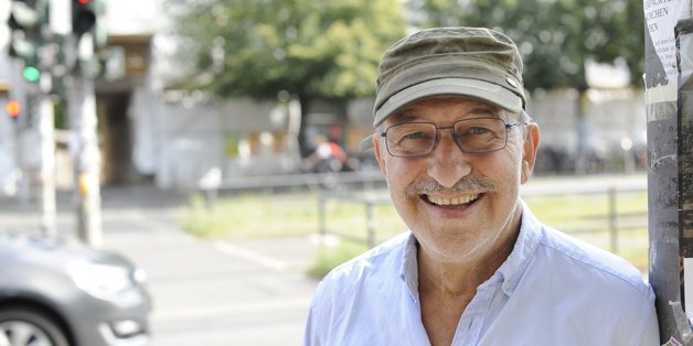 Ein älterer Mann mit einer grünen Schirmmütze lehnt an einer Straßenlaterne und lacht in die Kamera