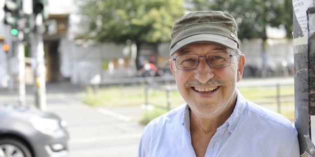 Ein älterer Mann mit einer grünen Schirmmütze lehnt an einer Straßenlaterne und lacht in die Kamera