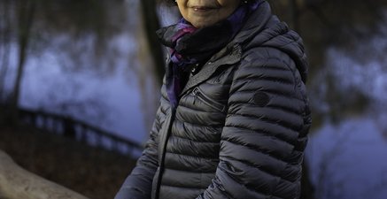 Eine ältere Frau lehnt an einem Holzgeländer, dahinter sind Bäume und ein Kanal zu sehen. Sie blickt in die Kamera.