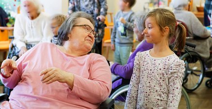 Eine Pflegeheimbewohnerin im Rollstuhl und ein Kita-Kind lächeln sich an.