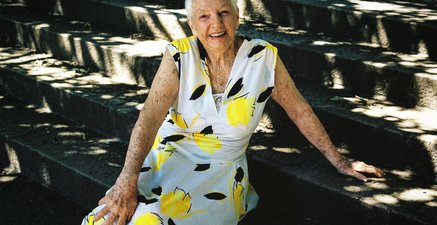 Maureen, eine 93-jährige Frau, trägt ein in strahlendem Gelb und Weiß gemustertes Sommerkleid und sitzt auf einer T