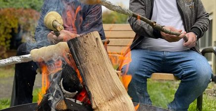 Zwei Männer sitzen an der Feuerschale und halten Stöcke mit Stockbrot ins Feuer. Sie wirken sehr fröhlich.