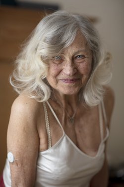 Ältere Frau mit langen Haaren sitzt Modell im zartgelben seidigen Unterkleid und lächelt.