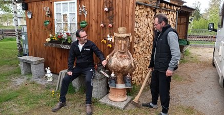 Ältere Zwillinge mit einer geschnitzten Holzfigur dazwischen. Dahinter ein Holzschuppen.