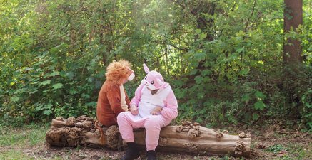 Senioren sitzen nebenaiander in Plüschtierkostümen auf liegendem Baumstamm. Löwe lacht Hase an.